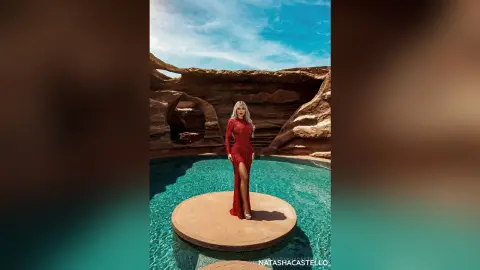 NatashaCastello poses in a long red dress on a stone platform in a desert pool under a blue sky.