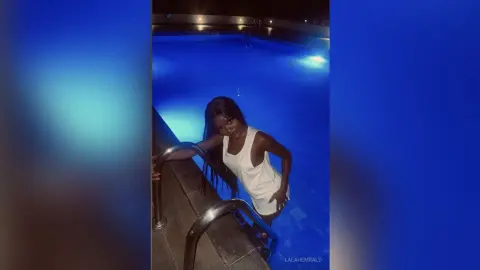 Young woman in a white sleeveless dress posing by a glowing blue swimming pool at night.