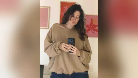 StellaNoire poses in a mirror wearing a tan oversized sweatshirt and dark jeans, looking down while holding her phone.