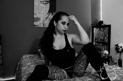 Victoria_Adams sits on a bed wearing a black tank top and fishnet stockings, looking toward the camera with long dark hair.
