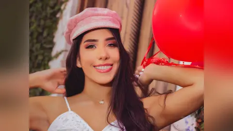 Smiling young woman in a white lace top and pink newsboy cap holding a red balloon outdoors.