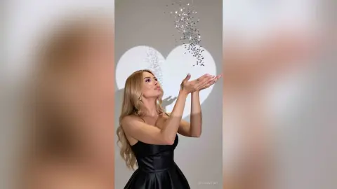 SerenaStar holds her hands up toward falling silver glitter while wearing a black satin dress.