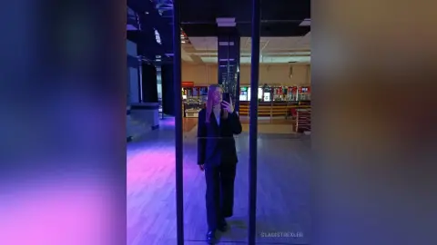 Stylish woman in a black blazer and trousers standing in a modern, moody glass hallway with purple lighting.