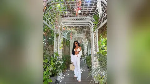 Stylish woman in a white crop top and trousers standing in a sunlit white garden pergola walkway.