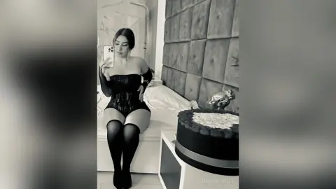 JessieOlivetti sits on a bed in a black lace bodysuit and long black stockings, taking a mirror selfie.