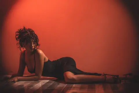 Moody portrait of a woman in a black dress reclining on a floor against a warm red background.