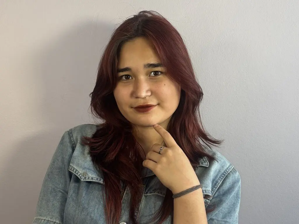 PatriciaGibson is a woman with long reddish-brown hair wearing a blue denim jacket, posing with her hand on her chin against a plain wall.