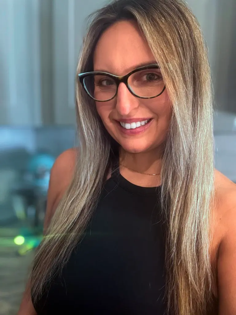 Smiling woman with long grey hair and glasses wearing a black tank top in a bright indoor setting.