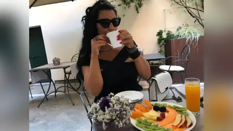 MissyJolie sips from a white cup while wearing black sunglasses and a black top at an outdoor table with fruit and juice.
