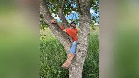 Cheerful woman in an orange shirt and blue jeans relaxing in a large tree in a sunny green meadow.