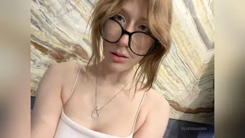 Blonde woman in a white camisole and black glasses posing against a marble wall background.