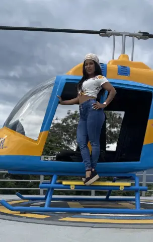 Stylish woman in white crop top and blue jeans posing by a yellow and blue helicopter under a cloudy sky.