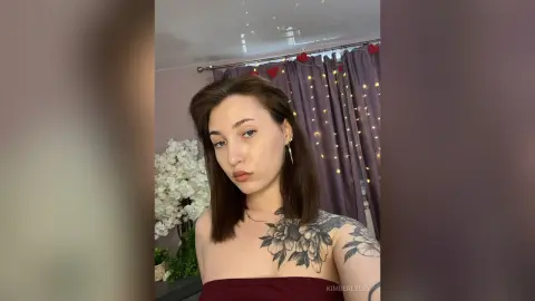 Young woman with tattoos in a burgundy top posing in a cozy room with fairy lights and white flowers.