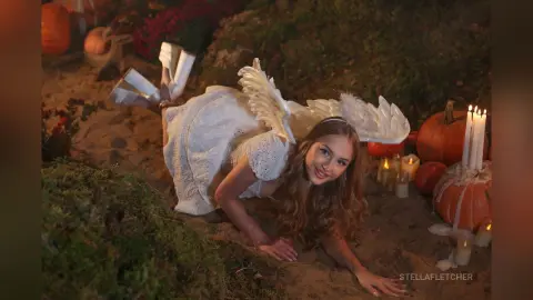 StellaFletcher lies on the sand in a white lace dress with white feathered wings, surrounded by pumpkins and lit candles.