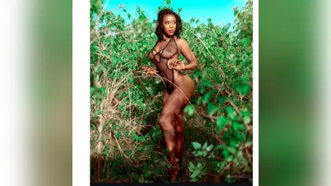 Confident Black woman in a black sheer bodysuit posing amidst lush green tropical foliage under a bright sky.