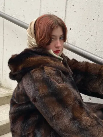 Redheaded woman in a luxurious brown faux fur coat sitting on concrete stairs in a moody urban setting.