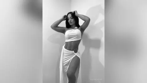 Elegant woman in a white halter crop top and flowing skirt posing against a minimalist wall in black and white.