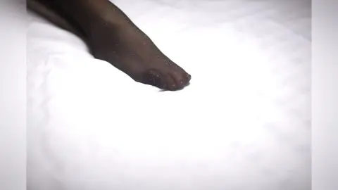 AryaSin shows off her legs and feet wearing sheer black stockings while lying on a white bed sheet.