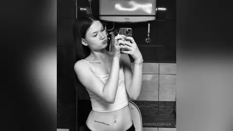 Young woman in a white tank top taking a playful mirror selfie in a modern bathroom.