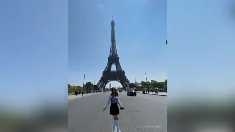 Stylish woman in a black sleeveless top and skirt walking near the Eiffel Tower in Paris under a clear blue sky.