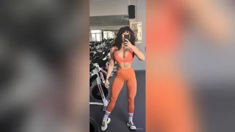 Fit woman in orange gym set taking a mirror selfie in a modern fitness center.