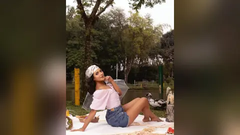 Smiling woman in a pink top and denim shorts relaxing on a white picnic blanket in a lush green park.