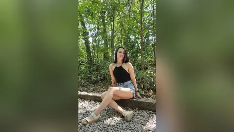 RossiNoleen sits on a wooden log in a lush green forest wearing a black halter top and denim shorts.