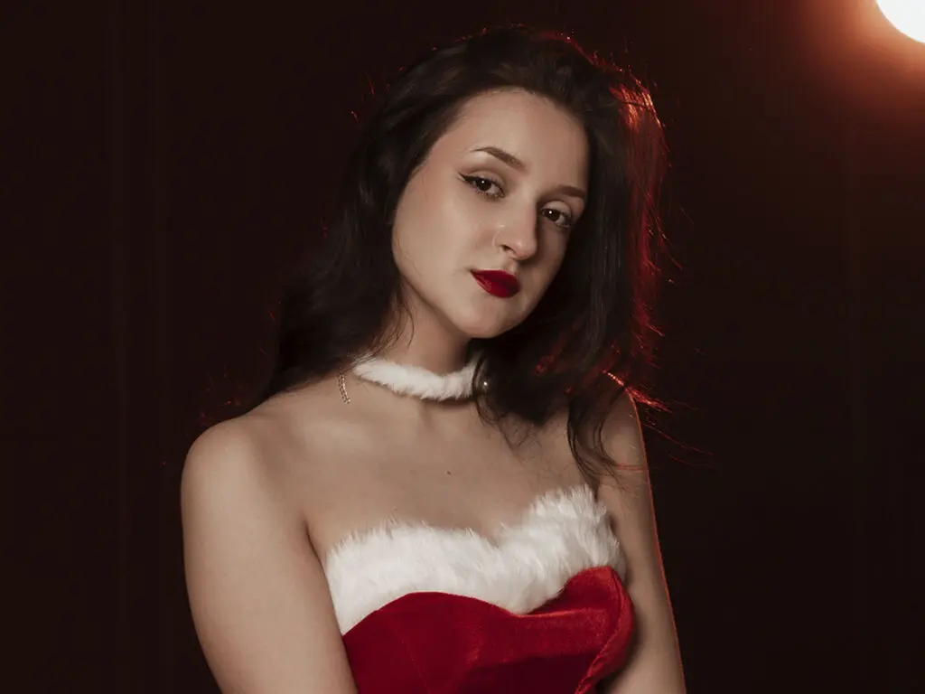 Young woman in a red and white velvet holiday outfit posing with a moody, elegant expression against a dark background.