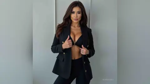 ReynaGomes poses in a black blazer and black bra, showing off her cleavage and midriff against a plain grey wall.