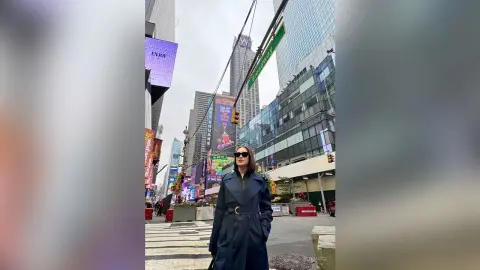 Stylish woman in a navy trench coat walking on a city street near Broadway skyscrapers under an overcast sky.