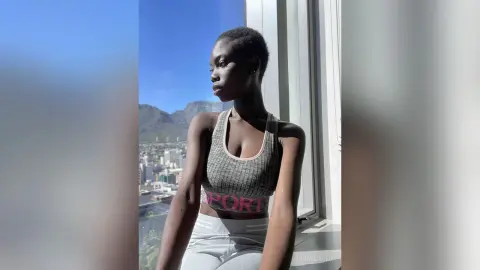 Young woman in grey sports bra posing by a window with a scenic mountain view during golden hour.