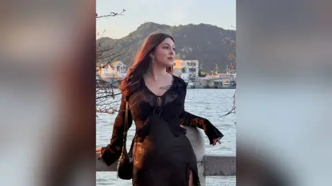 Young woman in a black velvet dress posing by a scenic lake with mountains at sunset.