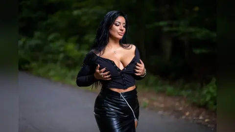 MissyJolie stands outdoors wearing a black long-sleeved top and a black leather skirt, using her hands to pull down her neckline and show cleavage.