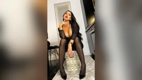 NicoleBella poses in a black sheer bodysuit, leaning forward over a stack of cash on a marble floor.