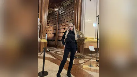 Elegant woman in black outfit standing in a grand vintage library with tall bookshelves and gold decor.