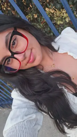 Smiling young woman with long dark hair wearing red glasses and a white blouse in a sunny garden setting.