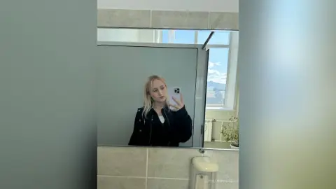 Blonde woman in a black jacket taking a mirror selfie in a bright, modern bathroom.