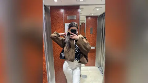 Woman in a brown jacket and white pants taking a mirror selfie in a modern elevator with marble walls.