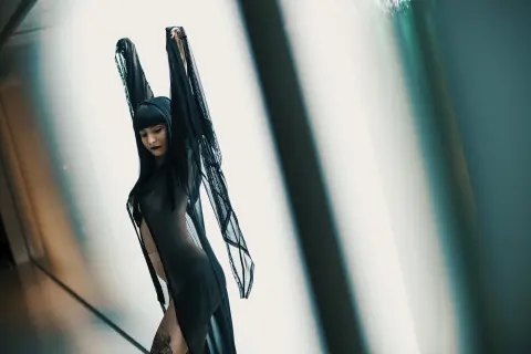 Moody portrait of a woman in a sheer black lace outfit standing against a bright, minimalist backdrop.