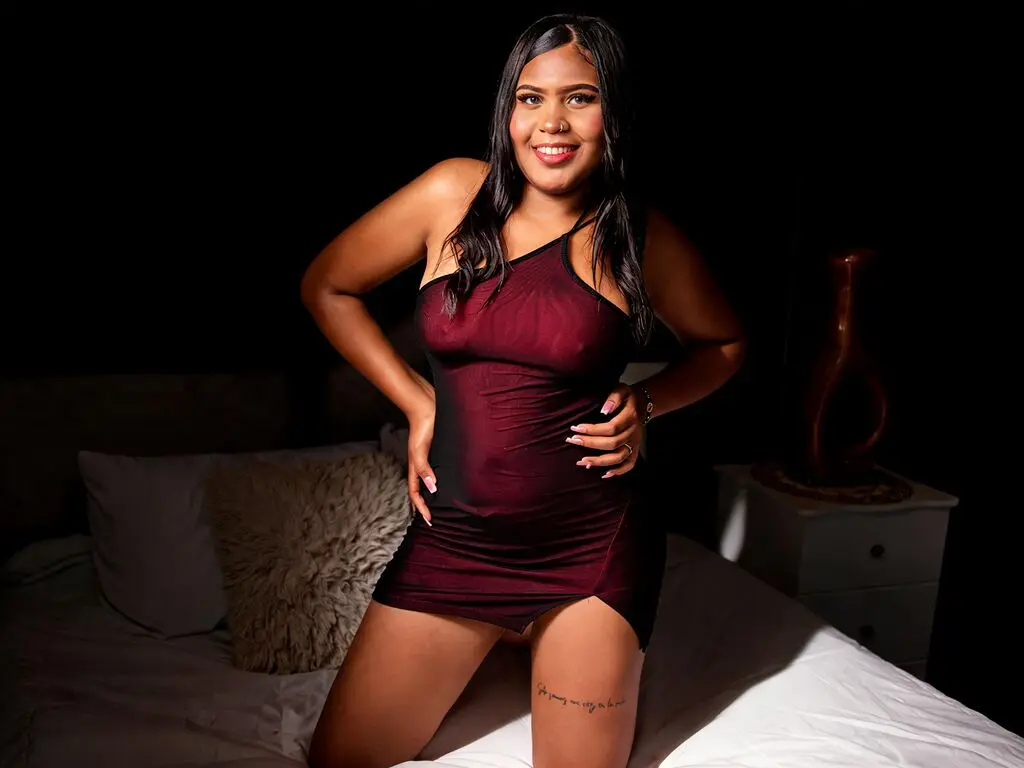 Smiling woman in a burgundy satin slip dress posing on a bed in moody low-light setting.