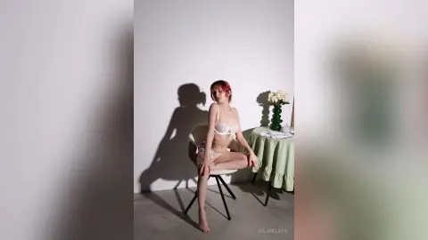 Young woman in white lace lingerie sitting on a chair in a minimalist, bright studio setting.