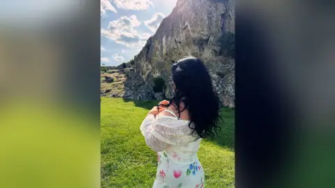 Woman in a white lace floral dress standing in a sunny green meadow near large rocky cliffs.