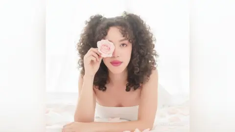 Curly-haired woman in a white tube top holding a pink rose against a bright, dreamy white background.