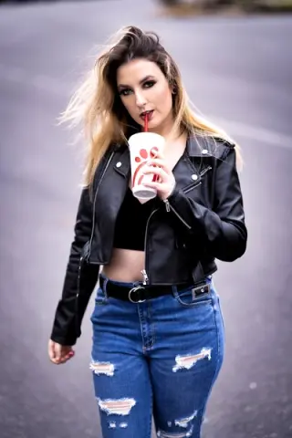 Young woman in a black leather jacket and ripped blue jeans sipping a drink on a street with a cool mood.
