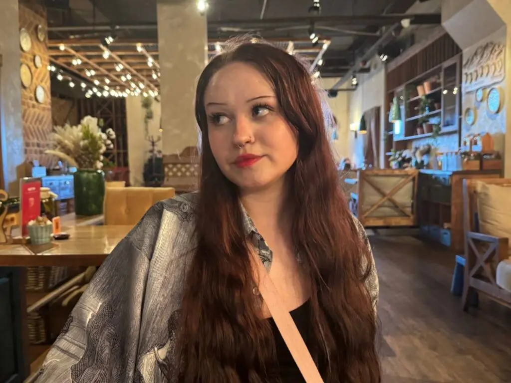 EmilyGreek poses with long wavy brown hair wearing a grey patterned shirt in a restaurant setting.