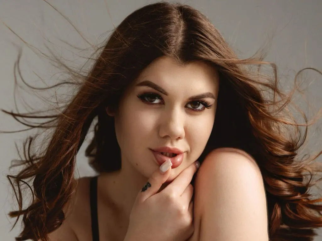 Beautiful woman with long flowing brown hair posing in a black top against a neutral studio background.