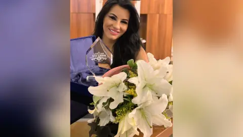 MissyJolie smiles while holding a clear award next to a large bouquet of white lilies.