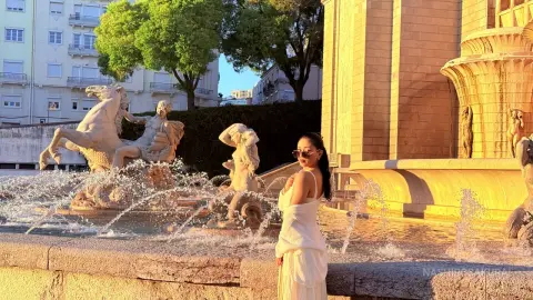 Young woman in a white summer outfit standing by a classical stone fountain in golden hour sunlight.