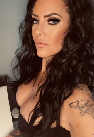 SensationalMya77 poses with long wavy dark hair and blue eyes, wearing a black lace top that shows off her cleavage and shoulder tattoo.