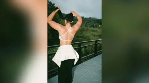 Athletic woman in a white bikini top and black pants tying her hair on a balcony overlooking a lush green forest.